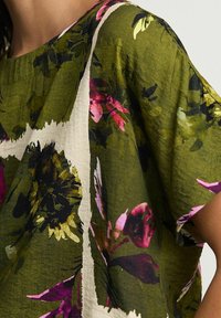 Bluzka w kwiatowy wzór w oliwkowym kolorze, z fioletowymi i różowymi akcentami, wykonana z lekkiego materiału, w luźnym kroju i z dekoltem łódkowym.