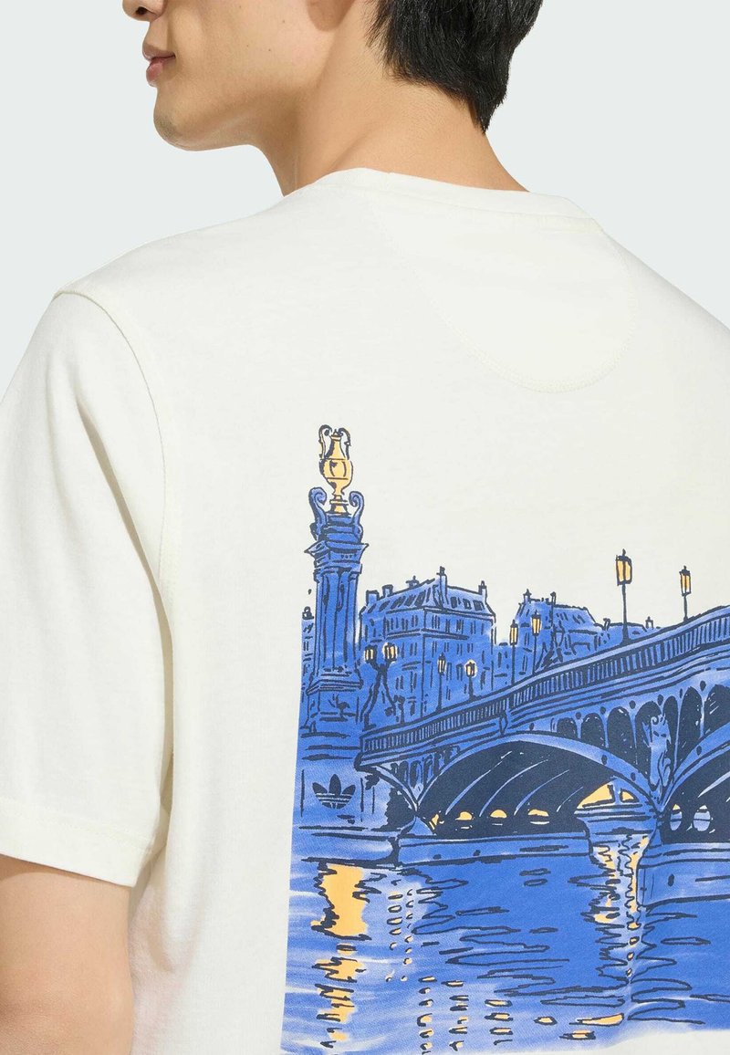 Gros plan d'une personne portant une chemise blanche avec une illustration bleue et jaune d'un pont de ville au-dessus de l'eau dans le dos.