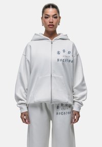 Felpa bianca con zip, dotata di una grande tasca frontale, vestibilità comoda e testo ricamato. Realizzata in tessuto morbido, presenta un cappuccio ampio.