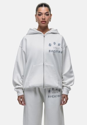 Felpa bianca con zip, dotata di una grande tasca frontale, vestibilità comoda e testo ricamato. Realizzata in tessuto morbido, presenta un cappuccio ampio.
