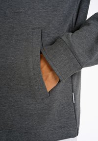 Grå fleece-tröja med sidoficka, ribbade ärmar och en texturerad tyg. Fickan har en kontrasterande brun inredning.