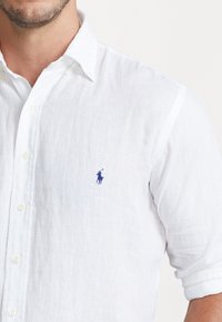 Vit linnen skjorta med button-down krage, korta ärmar och en blå broderad logotyp på vänster bröst. Lätt textur och andningsbart material.