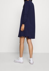 Sukienka w paski w kolorze granatowym z długim rękawem, o rozkloszowanym kroju i bocznych rozcięciach, zestawiona z białymi, wysokimi trampkami. Gładka faktura materiału.