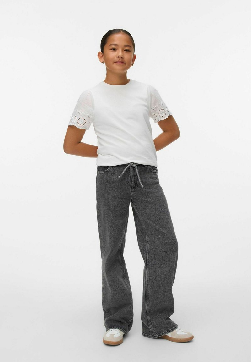 Niña joven de pie con confianza, vistiendo una blusa blanca de manga corta con bordados, jeans oscuros sueltos y zapatillas blancas.