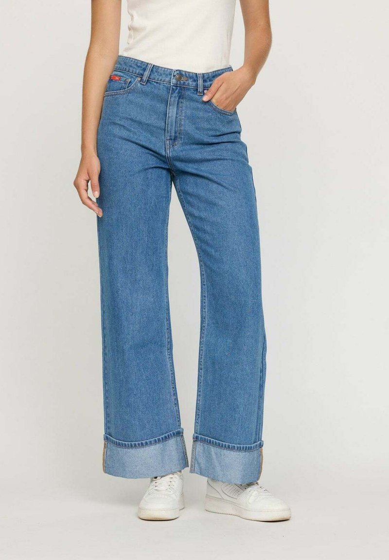 Femme portant un jean bleu taille haute à jambes larges avec des ourlets repliés et des baskets blanches, se tenant devant un fond uni.