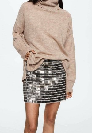 Femme portant un pull à col roulé beige oversized avec une main rentrée dans l'ourlet, associée à une mini-jupe argentée brillante et noire.