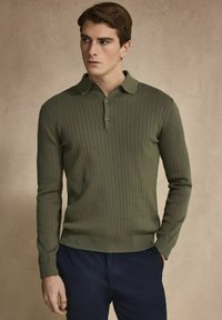 Olivegrön långärmad polo med ribbad textur, knäppbar krage och åtsittande ärmslut, kombinerad med marinblå byxor.