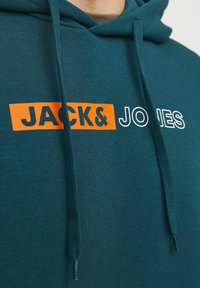 Sudadera de color verde azulado oscuro con un bolsillo tipo canguro, presenta el texto "JACK&" en naranja y "JONES" en blanco, cordones ajustables y tela texturizada.