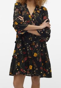 Svart blommig klänning med livfulla gula, röda och vita blommor, V-ringning, trekvartsärmar och ett lager-på-lager volangdesign.