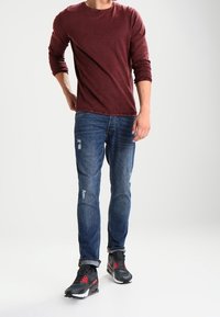 Chemise bordeaux à manches longues, jean bleu usé, et chaussures de sport noires avec des accents rouges. Coupe décontractée avec une texture douce.