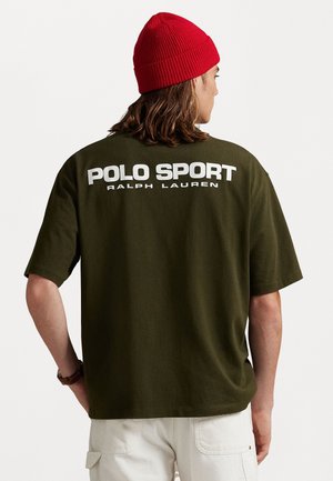 Person wearing an olive green oversized Polo Sport Ralph Lauren t-shirt and red knit beanie, facing away with hands behind back.