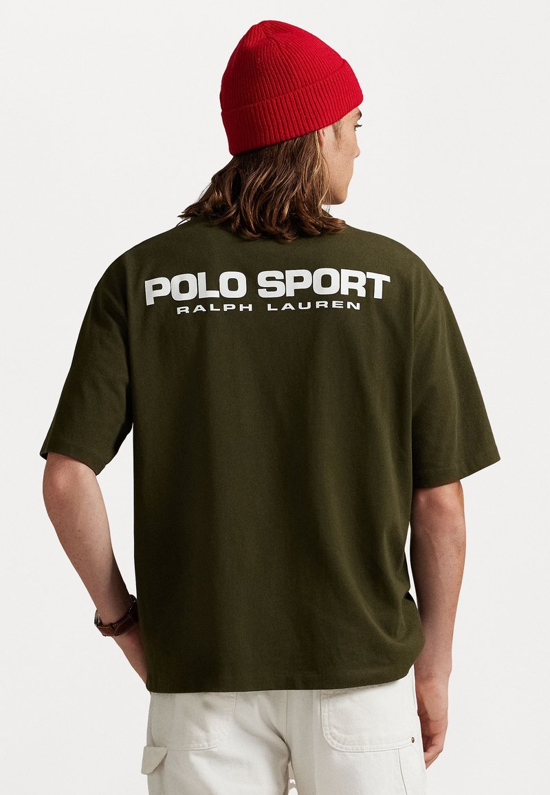 Olajzöld, oversized Polo Sport Ralph Lauren pólót és piros kötött sapkát viselő személy, háttal állva, kezei a háta mögött.