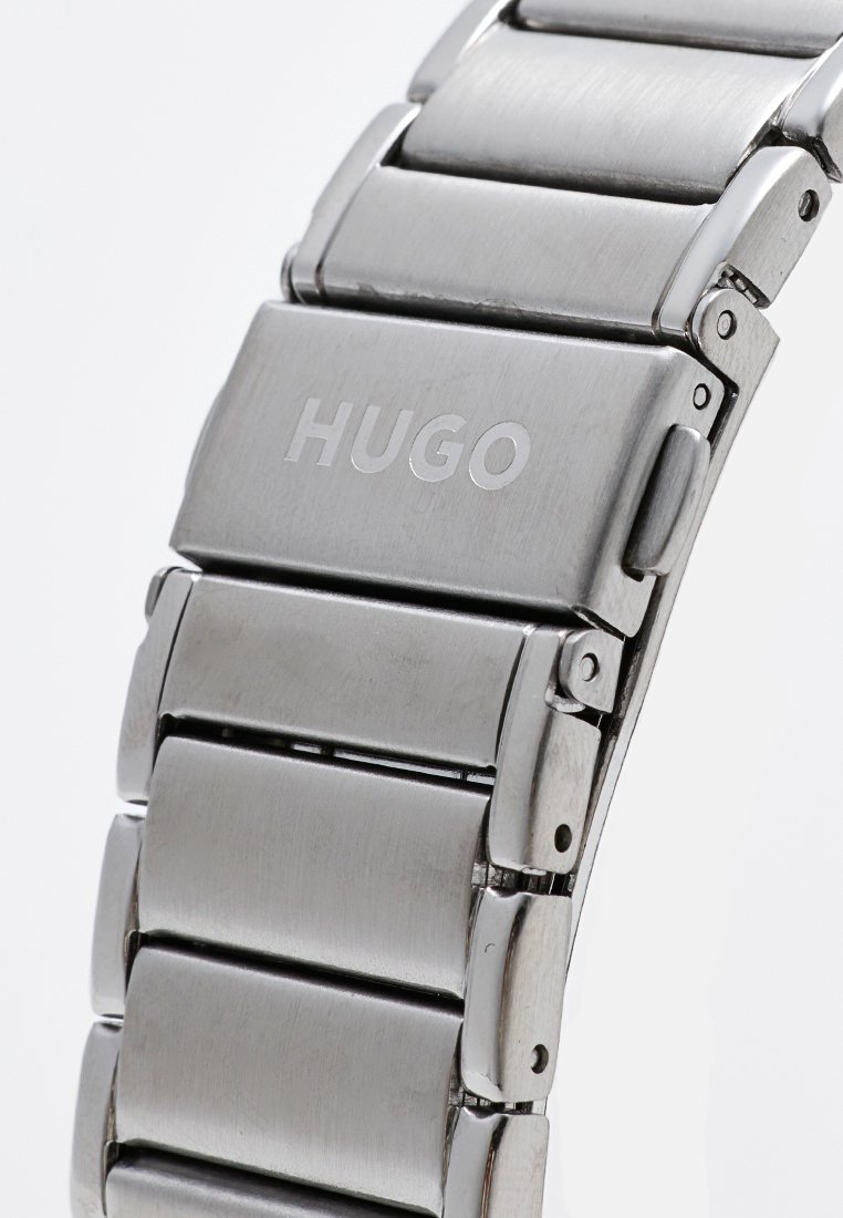 Pulsera de reloj de acero inoxidable con cierre desplegable grabado con el logo "HUGO", que muestra un acabado de metal pulido y cepillado.
