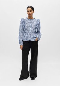 Femme portant un chemisier à volants bleu et blanc à motifs, un pantalon large noir et des chaussures noires, debout avec une main dans sa poche.