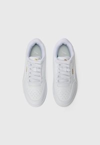 Et par hvite Puma-sneakers med snøring, perforert tåområde og gull Puma-logo, sett fra oven på en hvit bakgrunn.