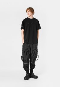 Camiseta negra con letras en relieve, combinada con pantalones cargo negros con múltiples bolsillos y cremalleras, y zapatos slip-on texturizados.