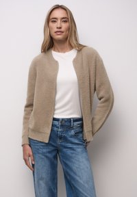 Cardigan beige texturé avec fermeture éclair à l'avant, encolure ronde et manches longues. Porté sur une chemise blanche, associé à un jean bleu clair.