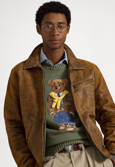 Καφέ δερμάτινο μπουφάν με ψηλό γιακά, πράσινο πλεκτό πουλόβερ με κεντημένη αρκούδα, φορεμένο πάνω από μπλε πουκάμισο με γιακά.
