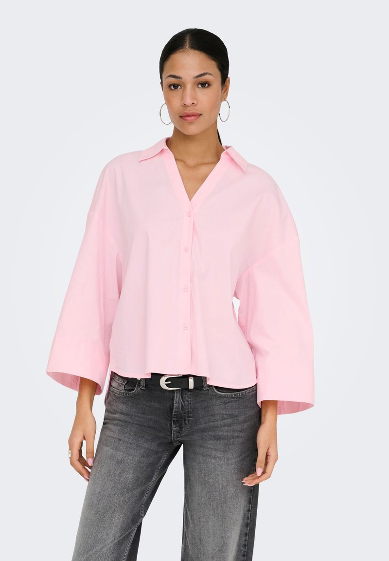Femme portant une chemise rose pâle surdimensionnée avec de larges manches et un jean noir délavé, sur fond uni.