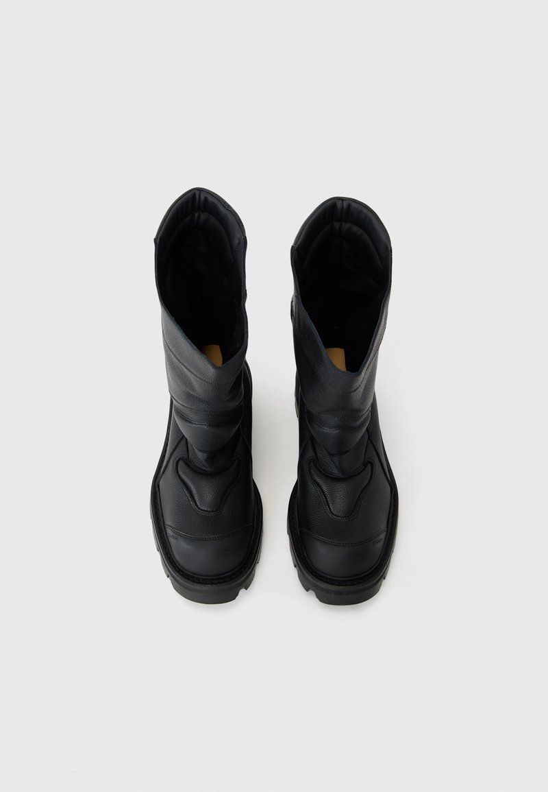 Bottines en cuir noir avec une finition texturée, présentant un design haut, des détails de couture et une forme arrondie. Doublure intérieure douce visible.