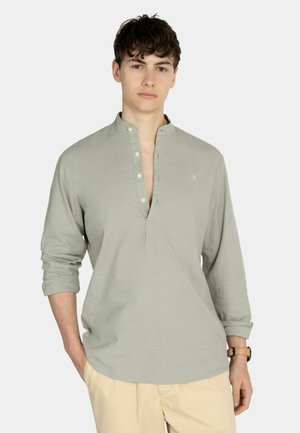 Joven con camisa de manga larga gris claro con cuello mandarín y pantalones beige sobre un fondo liso y claro.