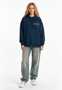 Námornícky modrá mikina s kapucňou a prednou vreckom, sťahovacou kapucňou a bielym vyšitým textom. Nosená s svetlomodrými roztrhnutými džínsami a bielymi teniskami.