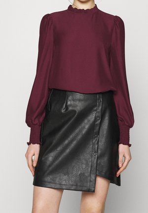 Mujer que lleva una blusa burdeos de manga larga con puños y cuello con volantes, combinada con una falda envolvente negra de polipiel con botones.