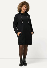 Mustapaita-mekko, jossa on pussi-kaulus, nyörikohdistukset, pitkät hihat, sivutaskut ja ribbitetyöt, yhdistettynä mustiin nilkkureihin.