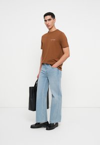 Καφέ βαμβακερό μπλουζάκι με λογότυπο, συνδυασμένο με ανοιχτόχρωμα φαρδιά τζιν παντελόνια, μαύρα παπούτσια και μαύρη τσάντα tote. Μινιμαλιστικός σχεδιασμός και υφές.