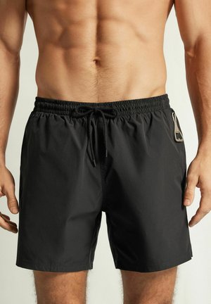 Męski tors w czarnych kąpielówkach z troczkiem w pasie i metalowym karabińczykiem przymocowanym do lewej bocznej kieszeni.