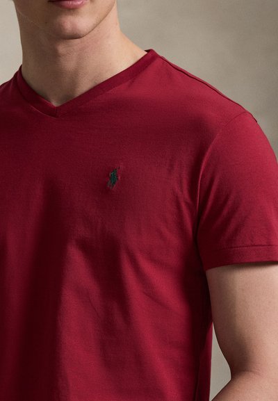 Άνδρας που φοράει εφαρμοστό κόκκινο T-shirt με κοντά μανίκια και V-λαιμόκοψη, με ένα μικρό σκούρο κεντητό λογότυπο αλόγου στο στήθος.