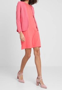 Kobieta idąca w koralowej sukience do kolan, teksturowanym różowym żakiecie i różowych butach na słupku z paskami na kostce, na jednolitym tle.