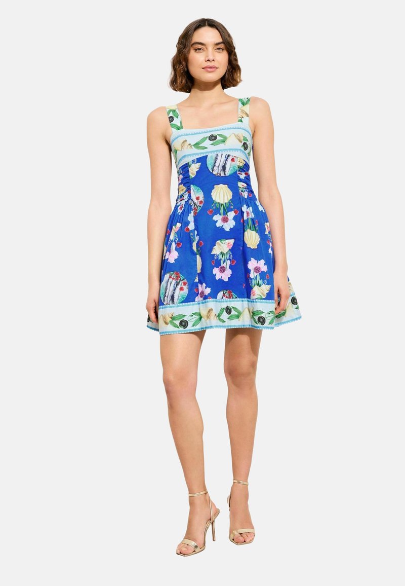 Woman wearing a blue sleeveless mini dress with floral and seashell patterns, standing with gold high-heeled sandals against plain background.