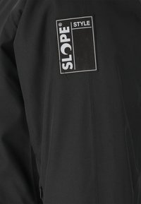 Musta takki, jossa on näkyvä suorakulmainen logopäällinen, jossa lukee "SLOPE STYLE." Kangas näyttää sileältä ja siinä on matta viimeistely.