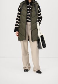 Kobieta ubrana w beżowe spodnie, czarno-biały sweter w paski, oliwkową puchową kamizelkę, czarne buty, trzyma czarno-białą torbę typu tote.