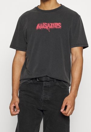 Mężczyzna noszący wyblakły czarny t-shirt z czerwonymi nadrukami oraz czarne dżinsy z czarnym paskiem, stojący na neutralnym tle.