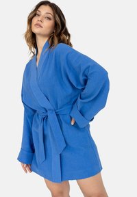 Niebieska lniana sukienka w stylu wrap z głębokim dekoltem w kształcie V, długimi rękawami, paskiem w talii i luźnym krojem, prezentująca teksturowany, lekki materiał.