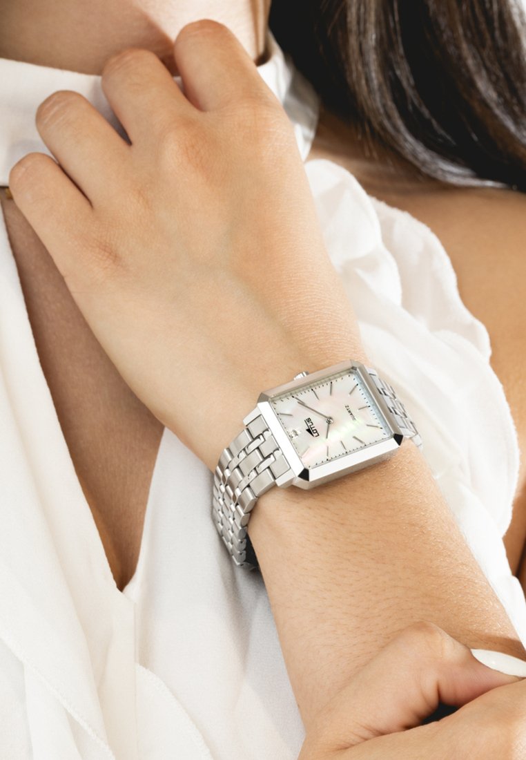 Reloj pulsera de metal plateado con caja cuadrada, esfera de nácar blanco y marcadores de hora plateados. Presenta una caja de metal pulido.