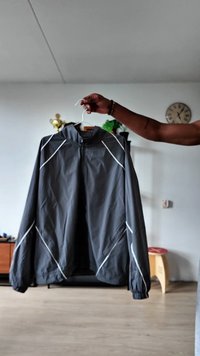 Hand holding a black jacket with white reflective stripes on a hanger inside a living room with a clock and plants.