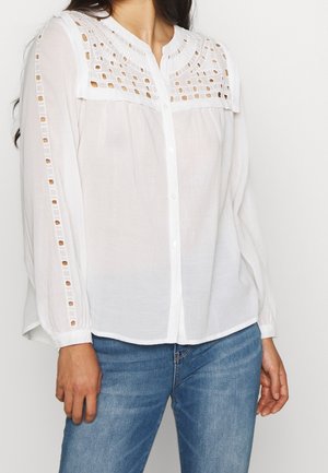 Hvid skjorte med knapper, der har en struktureret broderet yoke og udskæringer langs ærmerne, parret med blå denim jeans.