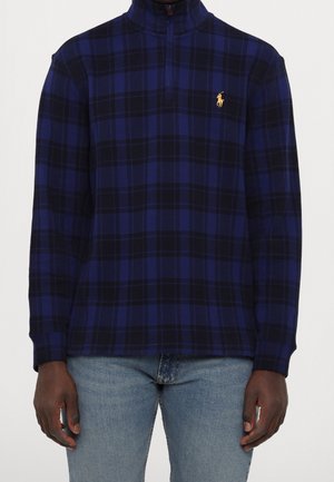 Polaire à fermeture éclair au quart en tartan bleu foncé et noir avec un petit logo brodé jaune sur le côté gauche de la poitrine, portée avec un jean bleu clair.
