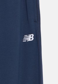 Jūras zilas sporta bikses, kas izgatavotas no gludas auduma, ar nelielu baltu izšūtu logotipu kreisajā pusē un taisnu griezumu.