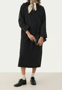 Czarna dżinsowa sukienka z długimi rękawami, kołnierzykiem mandarynkowym i przednią kieszenią, zestawiona z beżowym szalikiem i czarnymi skórzanymi butami.