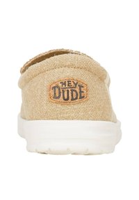 Zapato slip-on de lona beige con superficie texturizada, que presenta una etiqueta de cuero marrón que dice "HEY DUDE" y una suela de goma blanca.