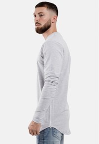 Grauhemd mit langen Ärmeln aus weichem Material, ausgestattet mit einem abgerundeten Saum und seitlichen Reißverschlüssen für zusätzliche Details, kombiniert mit hellblauen Jeans.