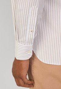 Chemise blanche à manches longues avec des rayures verticales beige clair, dotée d'un poignet à boutons avec un bouton accentué en rouge et d'un détail de poche pointue.