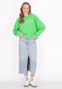 Sweatshirt verde con testo "Felicità Invisibile", abbinato a una gonna di jeans leggera, alta in vita, di colore blu chiaro, con uno spacco frontale. Sneakers bianche completano il look.