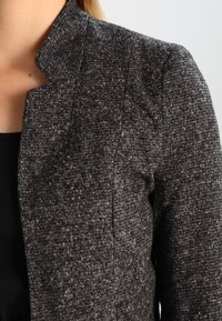 Gros plan sur un blazer gris foncé texturé porté sur un haut noir, montrant le col et la partie supérieure de la manche.