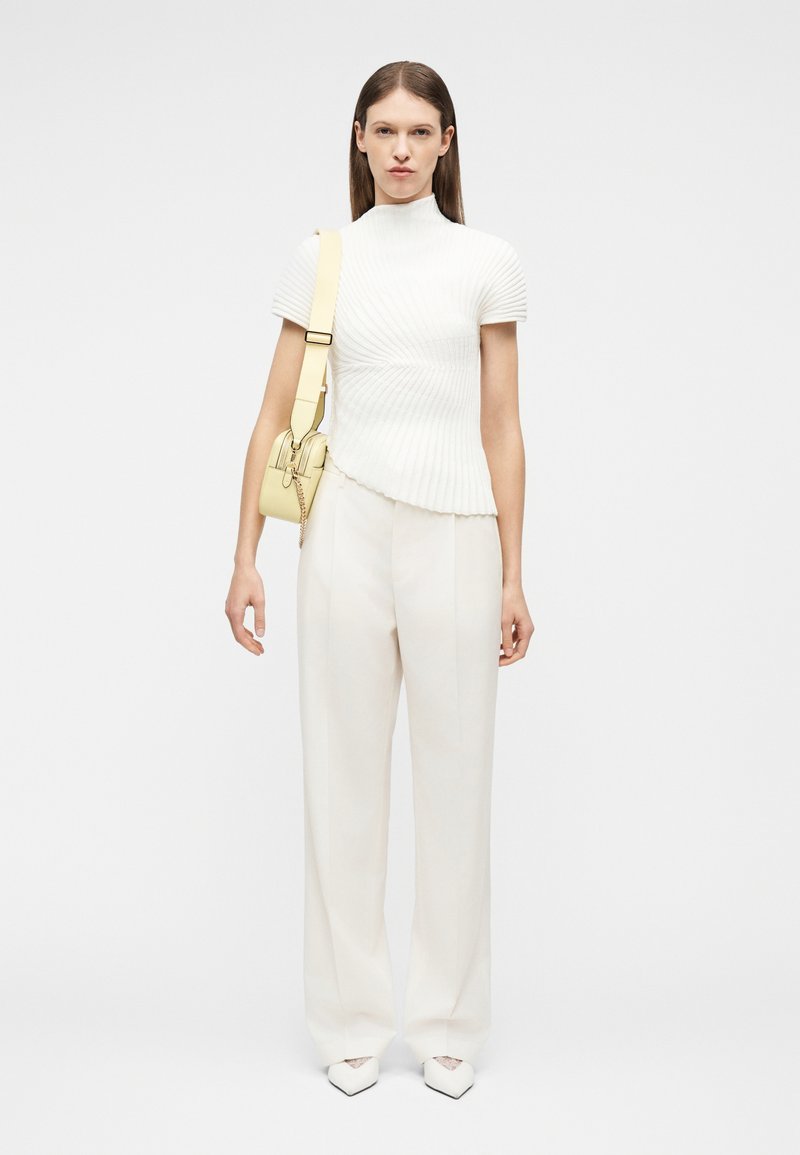 Γυναίκα που φοράει λευκό υφασμάτινο τοπ με κοντά μανίκια, λευκό παντελόνι με φαρδιά πόδια, λευκά μυτερά παπούτσια και μπεζ τσάντα ώμου με ιμάντα.