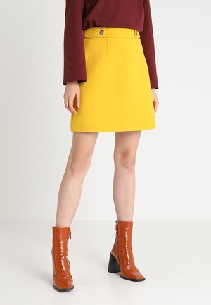 Żółta spódnica w kształcie litery A z pasem w talii i detalem guzika, zestawiona z błyszczącymi brązowymi botkami do kostki z blokowym obcasem i zamkiem.